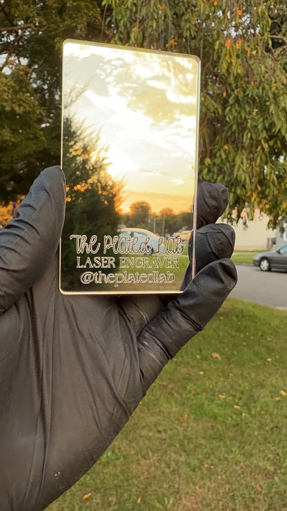 Gold mirror acrylic business card engraved with The Plated Lab logo, held up outdoors